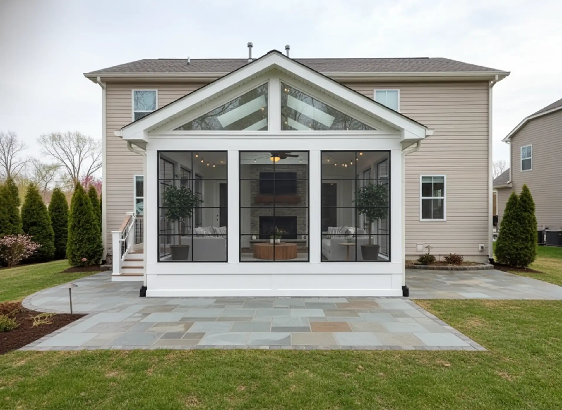 4 Season Sunroom Addition Ideas Modern 4-season sunroom addition featuring black-framed windows, a vaulted glass ceiling, an interior fireplace, and comfortable seating, built onto the back of a beige house with a stone patio.