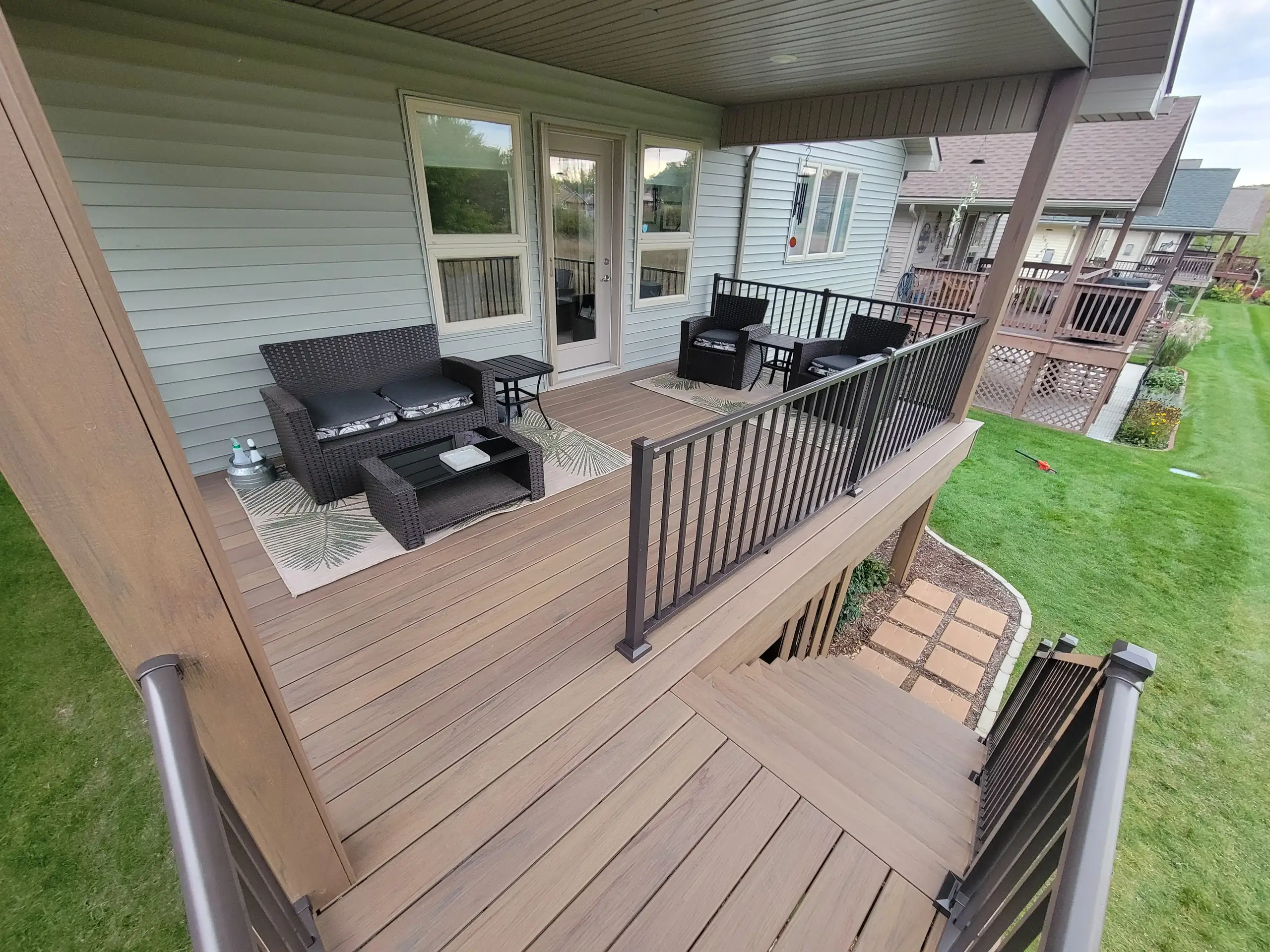Covered elevated composite deck with metal railing, wicker patio furniture, area rugs and stairs to a manicured lawn — composite decking vs wood decking