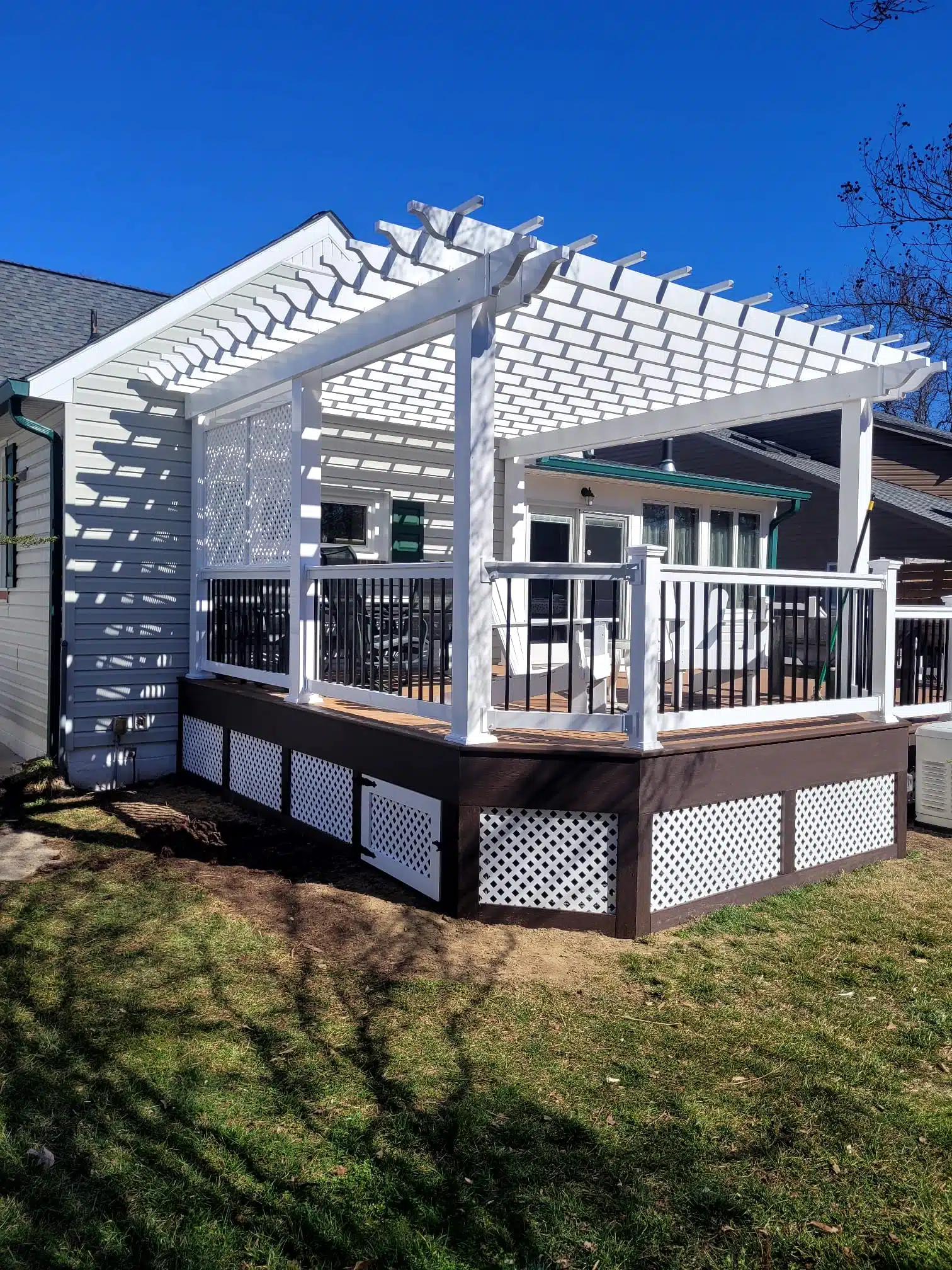 A white wood deck attached to a house. The deck has a white lattice skirt around the base and a white pergola-style roof structure. The railing is a combination of white top and bottom rails with black spindles. The house has light-colored siding, and the deck is situated over a grassy lawn