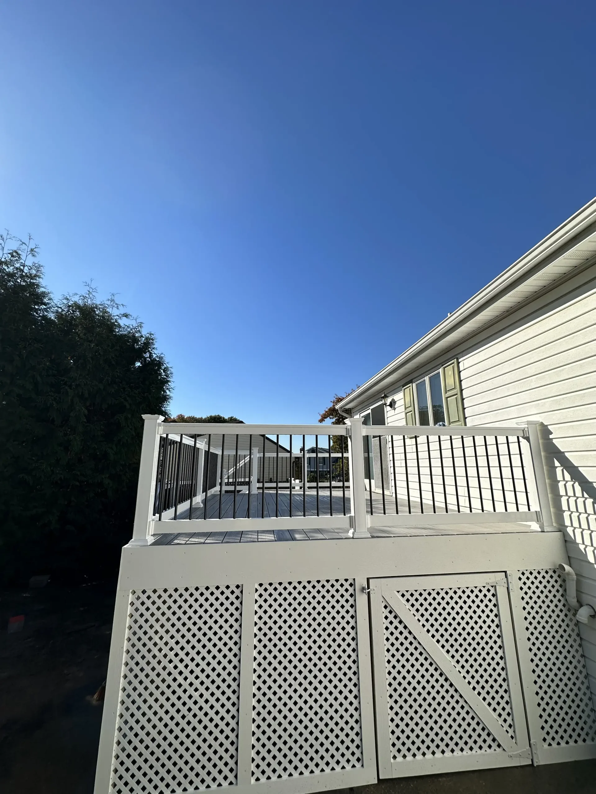 A white house exterior is shown, featuring white horizontal vinyl siding and a white gutter along the roofline. Below the siding, there is a small deck or porch. The deck has a black railing and white framing. Below the deck, there is white lattice skirting with two door-like panels.